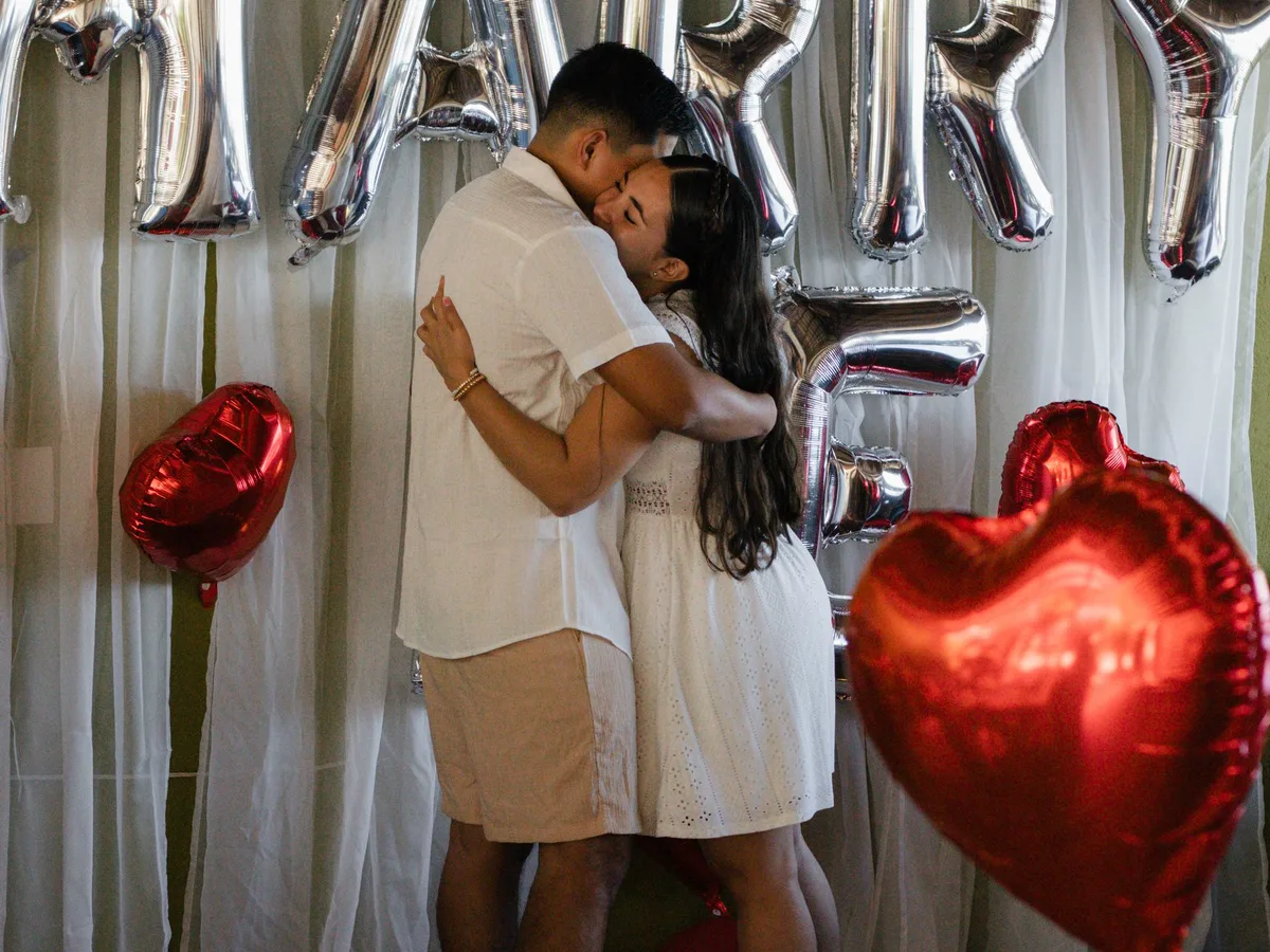 A couple embracing in front of MARRY ME foil balloons and red heart balloons
